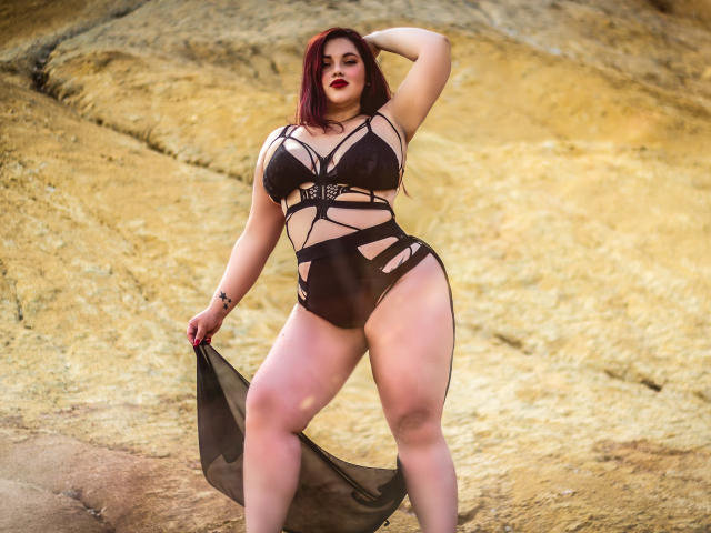 LissaClaire with red hair, curvy, large tits, wearing black strappy lingerie, holding sheer fabric, standing on rocky ground, looking confident, hand on head, red lipstick.