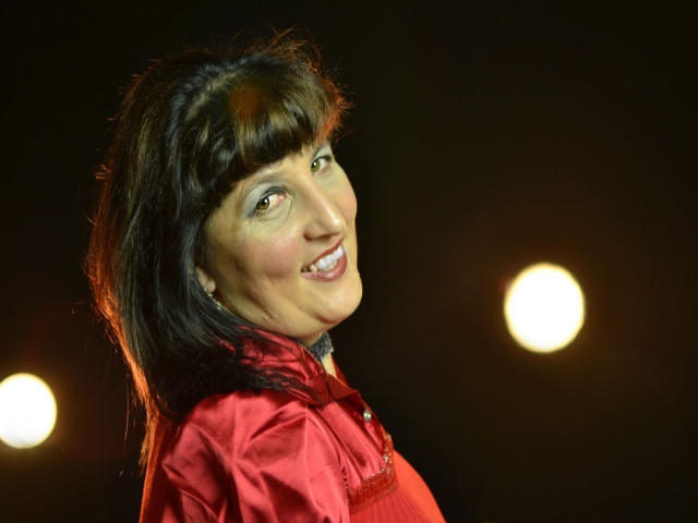 Ridemeraf, con cabello castaño oscuro y maquillaje azul en los ojos, sonriendo, vestida con una camisa de satén rojo, tiene senos de tamaño medio y está mirando por encima del hombro. Luces amarillas brillantes en el fondo..