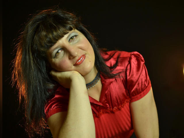 Ridemeraf, avec ses cheveux foncés et en désordre, son t-shirt rouge, ses yeux bleus et son rouge à lèvres rouge, la tête appuyée sur sa main, souriant légèrement, portant un collier noir, sur un fond sombre..
