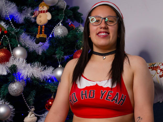 NatalieHarris with glasses, red cheerleader top, "NO HO YEAH" text, black choker, smiling, brown hair, Christmas tree with ornaments, and blue lights in the background.
