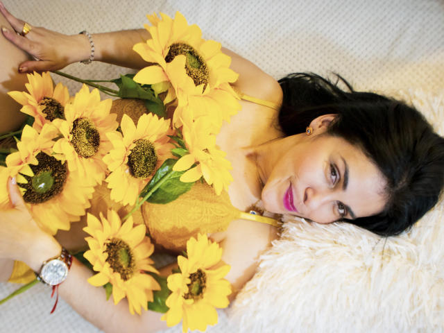 GirasolWalton with sunflowers, lying on a fluffy white pillow, wearing yellow lace, pink lipstick, black hair, and a silver watch, holding bright yellow sunflowers over her tits.