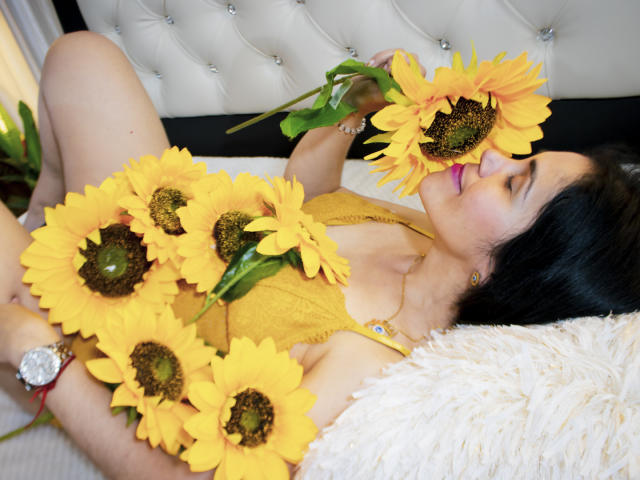 Girasol Walton ligt in bed, met zonnebloemen voor haar neus, gekleed in een gele kantjurk, met paarse lippenstift, en een horloge, haar zwarte haar is uitgespreid..