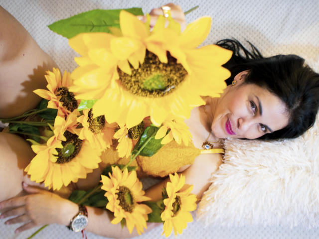 Girasol Walton con girasoles, acostada sobre un almohadón blanco y esponjoso, vestida con encaje amarillo, sonriendo, cabello oscuro, lápiz labial rosa, reloj en la muñeca, sosteniendo grandes girasoles amarillos, hojas verdes..