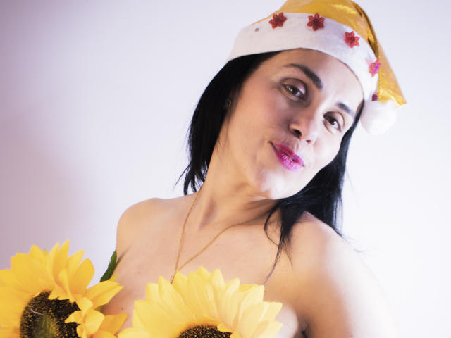 GirasolWalton with black hair, wearing a yellow and white Santa hat, holding sunflowers, has pink lipstick, and a gold necklace, smiling with pouty lips. Boobs visible.