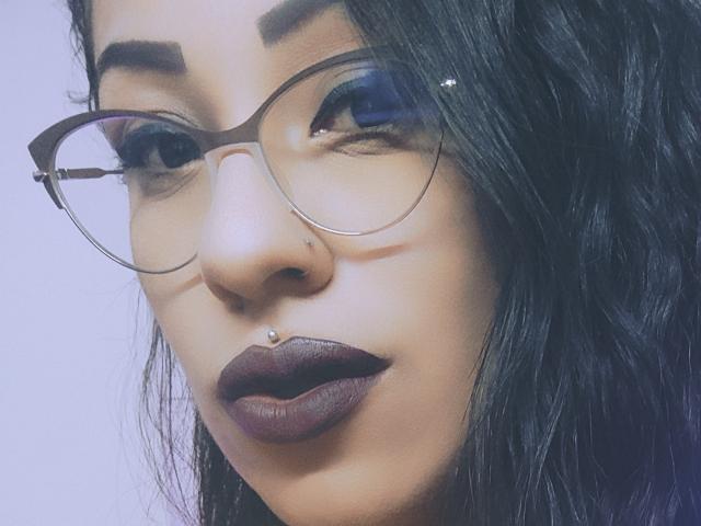 Le visage de LunaCandy, avec des lunettes, un rouge à lèvres violet foncé, des cheveux noirs ondulés, un piercing au nez et un piercing aux lèvres. Ses yeux sont ornés d'un fard à paupières bleu. Elle porte un petit anneau au nez..