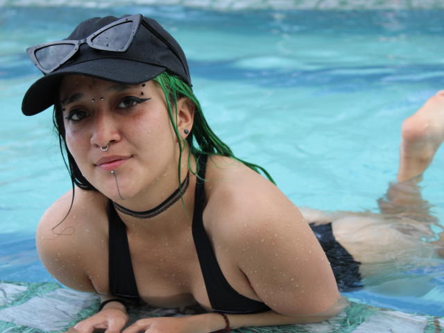 SalomePrince, com cabelo verde, boné preto e óculos de sol, deitada em uma piscina, usando um biquíni preto, mostrando seus seios e um piercing no nariz, com gotas de água na pele..