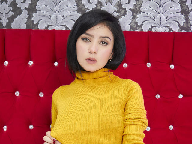 Kathy Rosse, com cabelo preto curto, vestindo um suéter amarelo de gola alta, um sofá vermelho com detalhes em botão e um papel de parede com padrão floral, tem uma expressão neutra e maquiagem discreta..