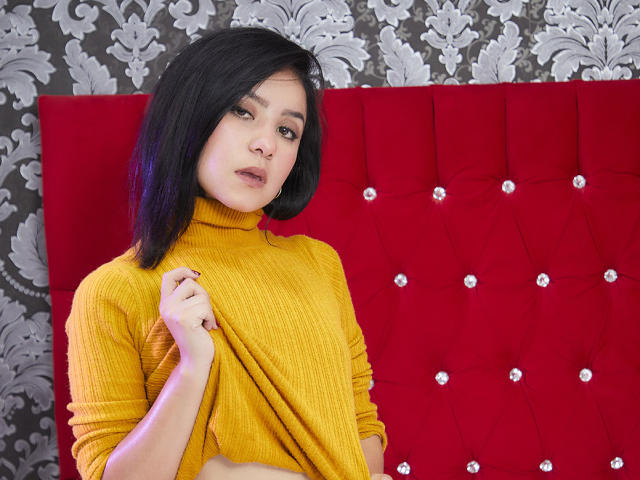 KathyRosse, avec des cheveux noirs courts, portant un pull jaune à col roulé, tenant le haut, regardant la caméra, mur rouge texturé en arrière-plan, papier peint à motifs..