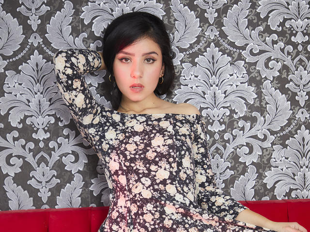 Kathy Rosse mit schwarzen Haaren, rosa Lidschatten und einem blumigen, schulterfreien Kleid, Arm erhoben, blickt in die Kamera, sitzt auf einem roten Sofa, trägt goldene Ohrringe im Stecker-Design, ernster Gesichtsausdruck..