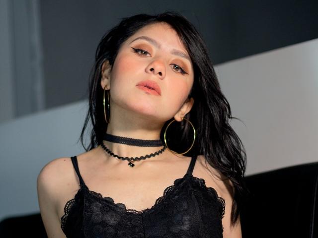 KathyRosse, com uma blusa preta de renda, brincos de argola verdes, um colar preto e cabelo preto ondulado, olhando para cima com os lábios rosados levemente entreabertos, maquiagem suave e um sorriso discreto..