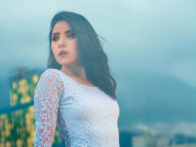 Kathy Rosse, con cabello negro ondulado, vestida con un vestido blanco de encaje, tiene senos pequeños y está mirando hacia un lado con una ligera sonrisa. El fondo está desenfocado..