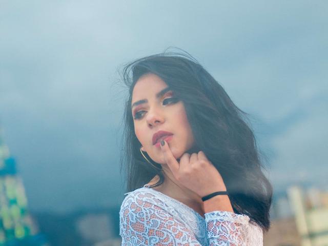 KathyRosse with long black hair, white lace top, finger touching her lip, red eyeshadow, gold hoop earrings, blue sky background, soft light, slight wind in her hair.
