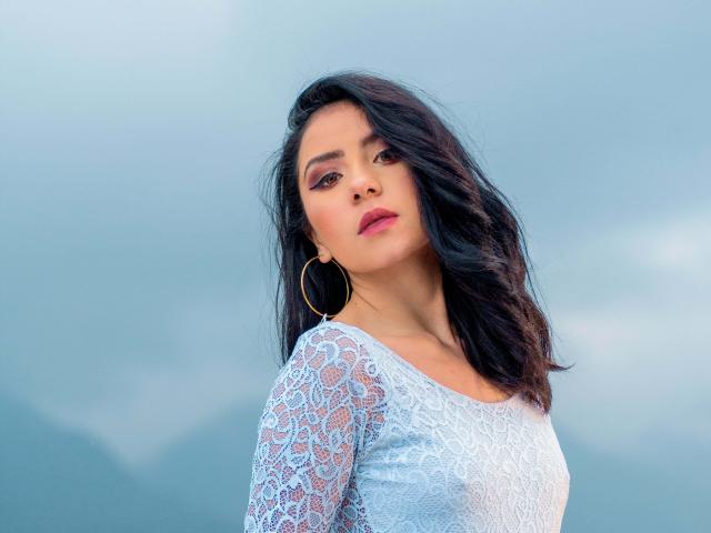 KathyRosse with wavy black hair, wearing a lace white top, gold hoop earrings, and pink lipstick, looking at the camera with a slight tilt to her head.