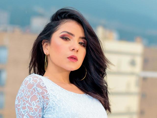 Kathy Rosse mit dunklen, welligen Haaren, leichtem Make-up, rosa Lippenstift, einem Spitzenoberteil, goldenen Reifenohrringen und einem ernsten Gesichtsausdruck. Ihre Brüste sind auf diesem Foto nicht sichtbar..