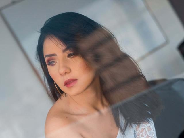 Kathy Rosse, con i capelli scuri, il rossetto rosso, gli orecchini a cerchio e la camicetta di pizzo bianco, appare pensierosa attraverso una finestra sfocata. Una luce soffusa mette in risalto la sua pelle chiara e i suoi tratti delicati..