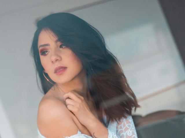 KathyRosse with dark, wavy hair, wearing a white, off-shoulder top, looking down with a slight frown, gold hoop earrings, soft pink lipstick, and subtle eye makeup.