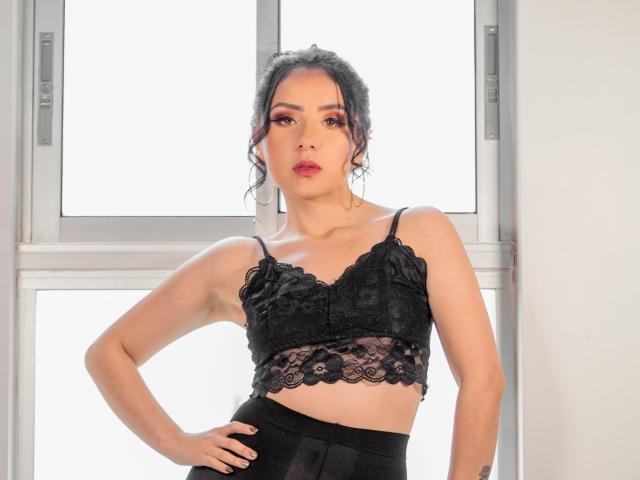 KathyRosse, avec un haut en dentelle noire, dévoilant de petites seins, un soutien-gorge en dentelle noire et une jupe noire, les mains sur les hanches, regardant l'appareil photo, une fenêtre en arrière-plan, peau pâle, rouge à lèvres rouge..