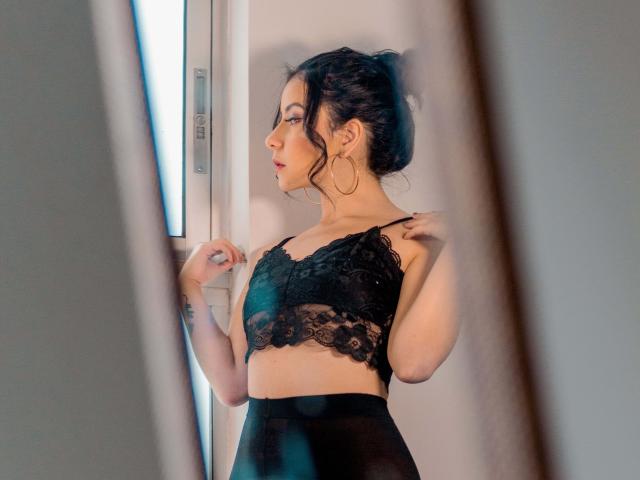 Kathy Rosse, con cabello oscuro recogido en un moño despeinado, vestida con un sujetador de encaje negro y pantalones negros de cintura alta, está de pie cerca de una ventana, mirando hacia afuera, sosteniendo la correa de su sujetador..
