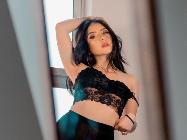 KathyRosse with long black hair, wearing a black lace bralette, has her arm raised, showing a tattoo, and looks seductively at the camera. Her tits are partially covered.