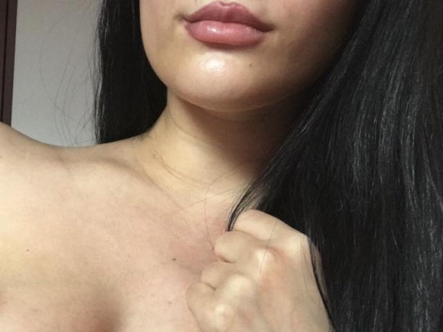 Ronina's glossy pink lips, slightly parted, with a small, shiny spot on her chin. She's holding a lock of her long, black hair, showing her bare shoulders.