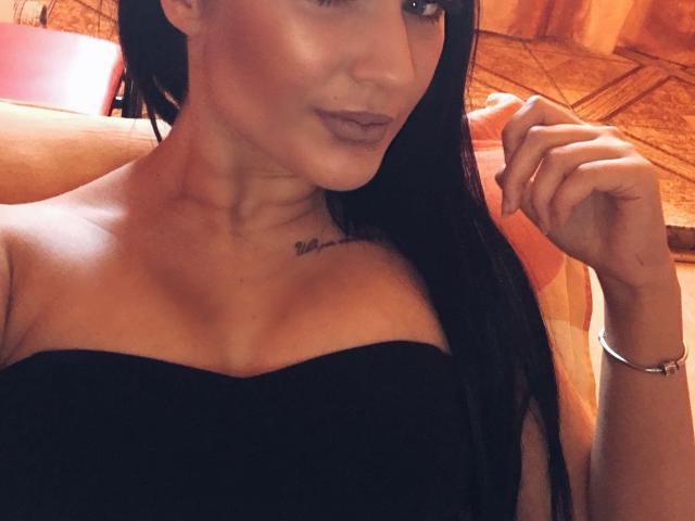 Ronina con lunghi capelli neri, seno di media grandezza, top nero senza spalline, tatuaggio sulla clavicola, sorriso appena accennato, occhi marroni, mano destra sollevata, braccialetto d'argento, luce calda, pelle leggermente lucida..
