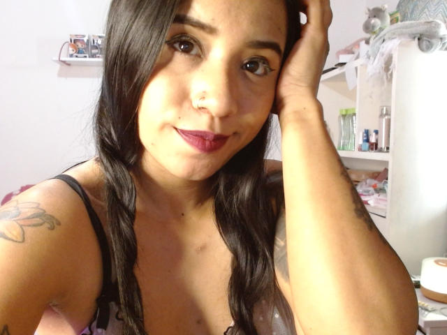 AshleyLover mit langem, schwarzem Haar, dunklen Augen und rotem Lippenstift, trägt ein schwarzes Oberteil, berührt ihren Kopf, zeigt ein Tattoo auf ihrem linken Arm und lächelt in die Kamera..