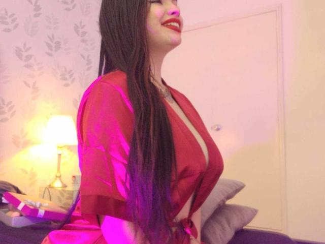 BrithneyLee mit langen braunen Haaren, rotem Lippenstift und einem roten Seidenbademantel, die große Brüste zeigt, sitzt auf einem Bett, lächelt, im Hintergrund ist ein violettes Licht, eine Lampe, ein Kissen und eine graue Wand zu sehen..