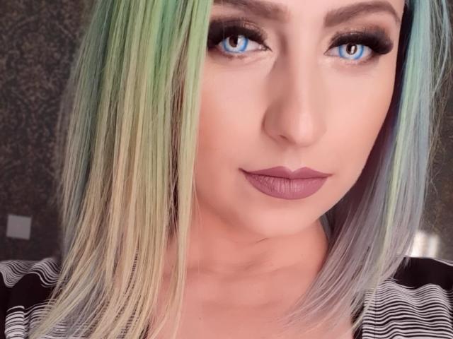 Agaa tem olhos azuis, cabelo que varia do verde ao loiro, e batom roxo. Sua maquiagem inclui delineador escuro e sobrancelhas grossas. Ela está vestindo uma camisa listrada..