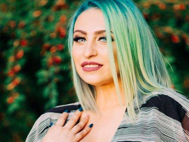 Agaa has green-blue hair, black and white striped shirt, and blue nails, smiling with pink lipstick, hand near chest, looking slightly to the side.