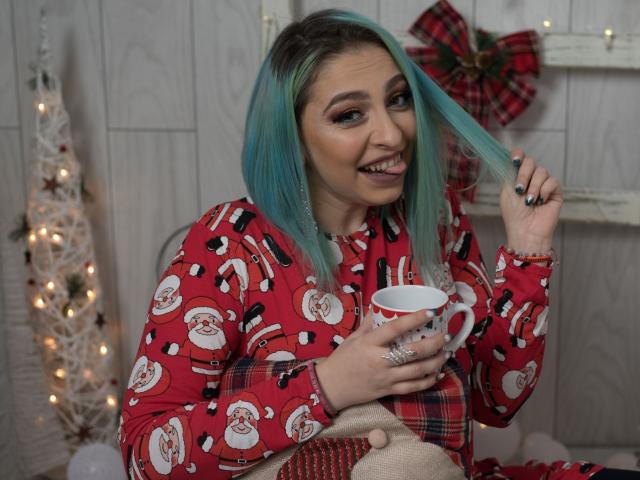 Agaa, com cabelo azul-esverdeado, vestindo um pijama de Natal vermelho, segurando uma caneca branca, sorrindo e puxando o cabelo de forma divertida com unhas pretas, mostrando os seios levemente por baixo do tecido..