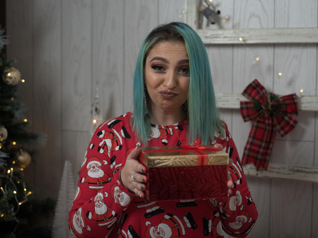 Agaa with teal hair, pouty lips, wearing red Christmas-patterned shirt, holding gold and red gift box, standing in front of decorated white wall, plaid bow on right.