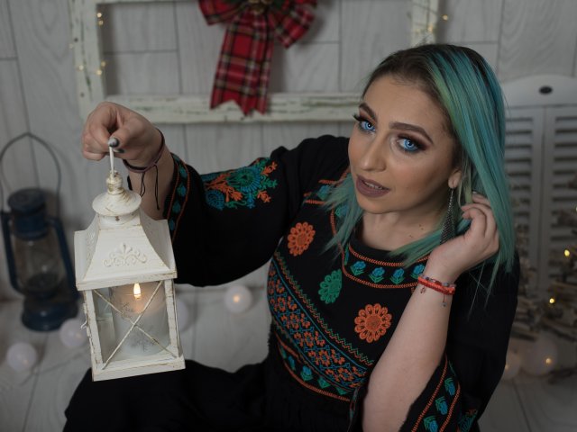 Agaa with teal hair holds a white lantern, wearing a black dress with colorful patterns, blue eyes, and dark lipstick. Her hand touches her hair, red bracelet visible.
