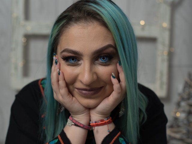 Agaa with blue-green hair, hands on cheeks, smiling at camera. She has blue eyes, dark makeup, and teal nail polish. Wearing black top with colorful bracelets.