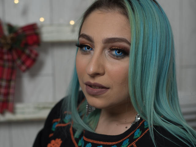 Agaa, com cabelo azul-turquesa, olhos azuis e maquiagem escura, exibindo seus lábios carnudos e nariz delicado, vestindo uma blusa preta com bordados coloridos. Suas nádegas não estão visíveis..