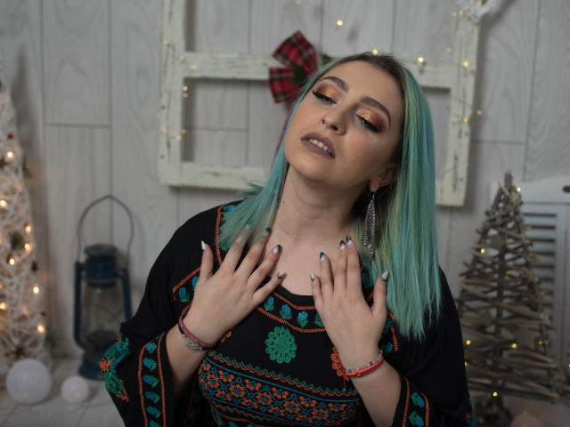Agaa, avec ses cheveux turquoise, les yeux fermés et un maquillage séduisant, porte une robe noire ornée de motifs colorés, les mains près de sa poitrine, le regard tourné vers le haut, et elle porte des boucles d'oreilles argentées ainsi que des bracelets colorés..