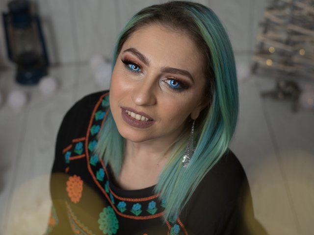 Agaa, com cabelo azul-esverdeado, olhos azuis e maquiagem escura, vestindo uma blusa preta com padrões coloridos, sorrindo para a câmera. Seus seios estão cobertos pela blusa..
