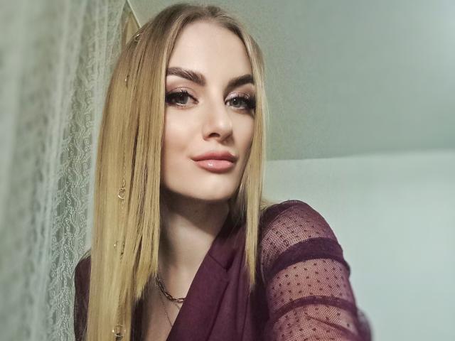 BlondyOneSex, met recht blond haar, draagt een doorschijnend donkerrode top, heeft volle lippen, lange wimpers en een zelfverzekerde glimlach, waarbij een subtiele gouden ketting te zien is..