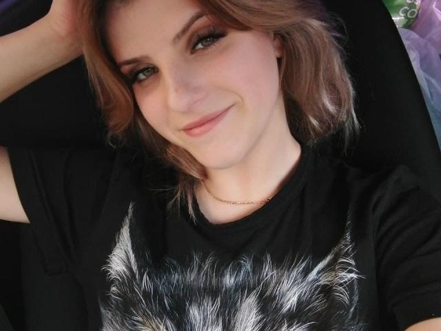 Alexandra Watson hat hellbraunes Haar, grüne Augen und ein süßes Lächeln. Sie trägt ein schwarzes Hemd mit einem fellartigen Muster und liegt mit einem Arm hinter dem Kopf..
