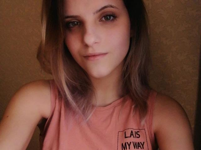 AlexandraWatson has light brown hair, green eyes, and a subtle smile. She's wearing a pink sleeveless top, showing her small tits. Her skin is fair and she's cute.