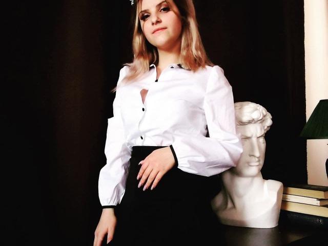 Alexandra Watson, con i capelli biondi, una camicia bianca con bottoni, una gonna nera, in piedi, con una mano appoggiata al fianco, accanto a un busto di marmo bianco, una lampada verde e libri su un tavolo di legno scuro..
