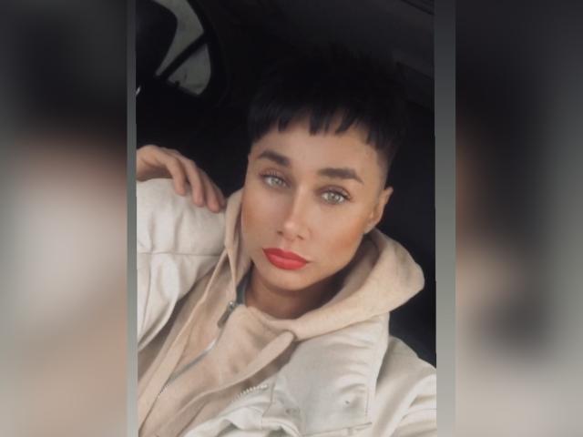 Ernesto Bergin, con i capelli neri corti, gli occhi verdi e il rossetto rosso, indossa una giacca beige con cappuccio e ha una mano sul collo, guardando nella fotocamera..