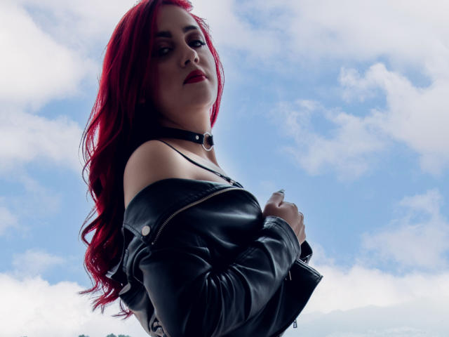 BrunaRousse69, avec des cheveux roux, une veste en cuir noir et un collier, tenant la veste, montrant ses seins, sur un ciel bleu avec des nuages, regardant vers le bas avec une expression confiante..