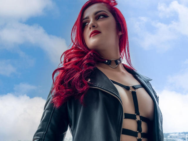 BrunaRousse69 met rood haar, een zwarte leren jas en een zwarte top met bandjes, die haar borsten laat zien, draagt een choker, kijkt zelfverzekerd omhoog, tegen een blauwe lucht met witte wolken..