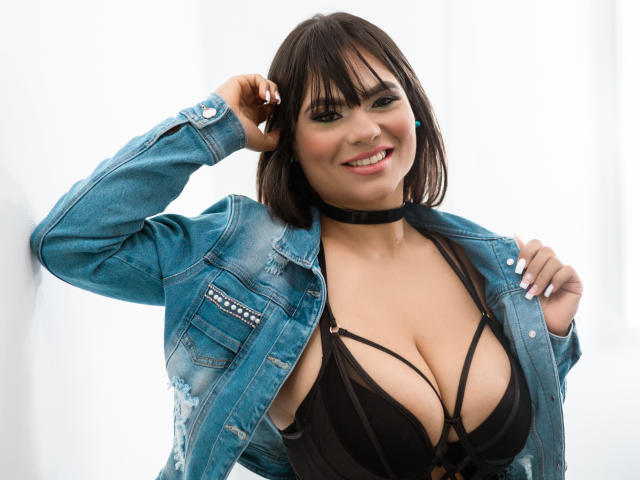 Briana Preston, con un corte de pelo bob castaño oscuro, sonriendo, vistiendo una chaqueta vaquera azul sobre un sujetador negro con tirantes, mostrando sus grandes senos, y mordisqueando un collar negro..