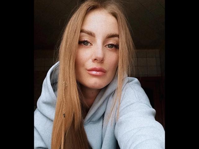 BlondyOneSex ha capelli lunghi e lisci, di colore castano chiaro, occhi verdi e labbra rosa lucide, indossa una felpa azzurra e ha una piccola forcina da capelli sul lato sinistro..