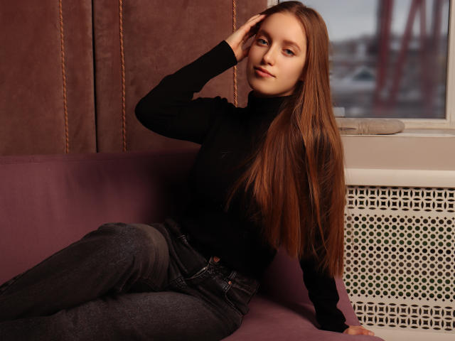 EvaPink, con lunghi capelli castani, un maglione nero a collo alto e jeans scuri, seduta su un divano viola, con una mano sulla testa, che guarda la fotocamera, luce soffusa, rilassata, sensuale..