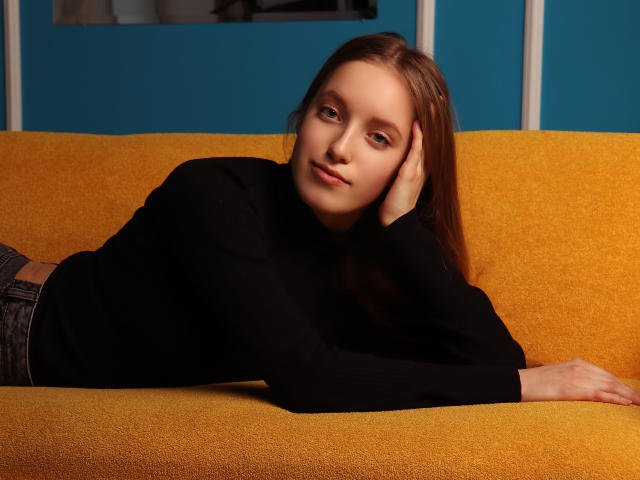 EvaPink, with light brown hair, black turtleneck, and gray pants, lays on a yellow couch, head on hand, looking relaxed and cute. Her ass is partially visible.