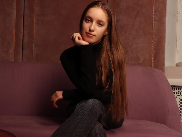 EvaPink with long brown hair, sitting on a purple couch, wearing a black turtleneck and gray pants, hand near mouth, looking at camera, soft smile, relaxed.