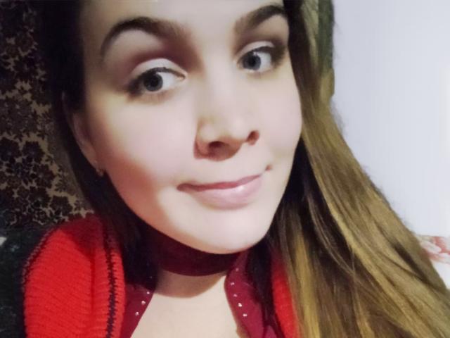 SindikaNiks with light skin, brown eyes, and long brown hair, wearing a red choker and red top, smiling subtly, showing a cute face and small nose.