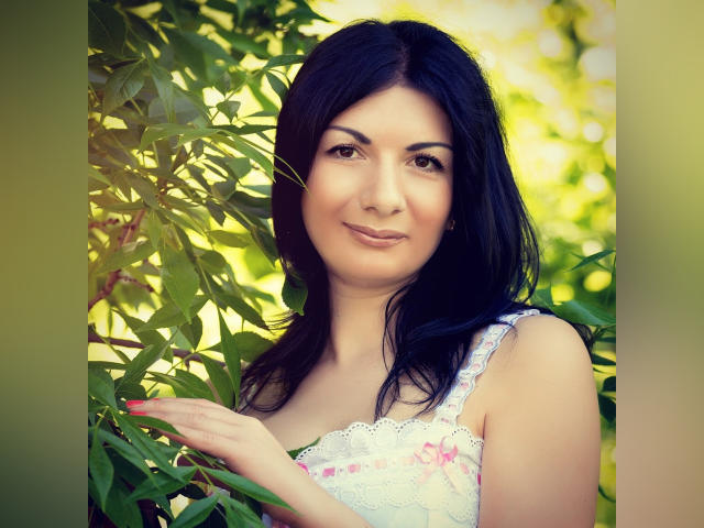 ZefirkaAxe, con cabello oscuro, sonriendo, de pie entre hojas verdes, vestida con un vestido blanco de encaje, con la mano tocando una hoja, luz suave, rostro bonito, linda, maquillaje sutil..